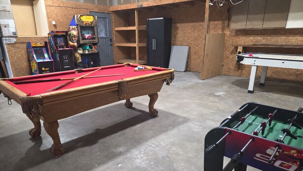 A pool table with a red felt surface, a foosball table, and arcade machines in a room with wooden walls and a concrete floor.