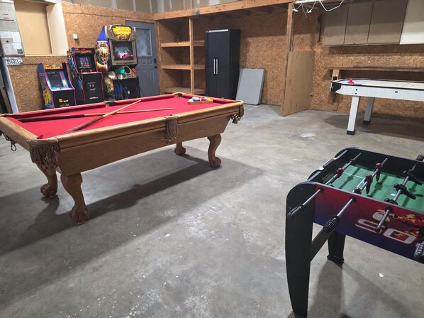 A pool table with a red felt surface, a foosball table, and arcade machines in a room with wooden walls and a concrete floor.
