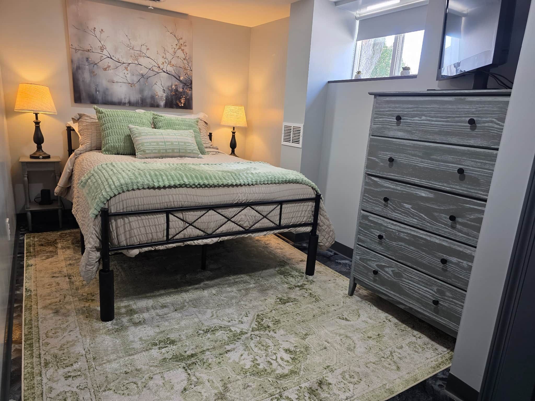 A bedroom with a bed, two bedside lamps, a dresser, and a large window.
