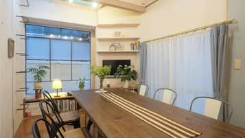 Un comedor con una mesa de madera, sillas, una estantería, una lámpara y un ventanal con cortinas.
