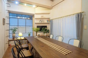 Un comedor con una mesa de madera, sillas, una estantería, una lámpara y un ventanal con cortinas.