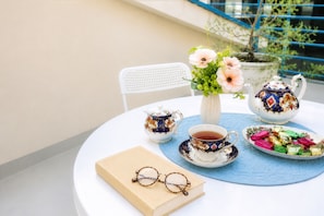 Una mesa redonda con una silla blanca, un libro, un par de gafas, un hervidor de agua, una taza, un plato de bombones y un jarrón con flores.