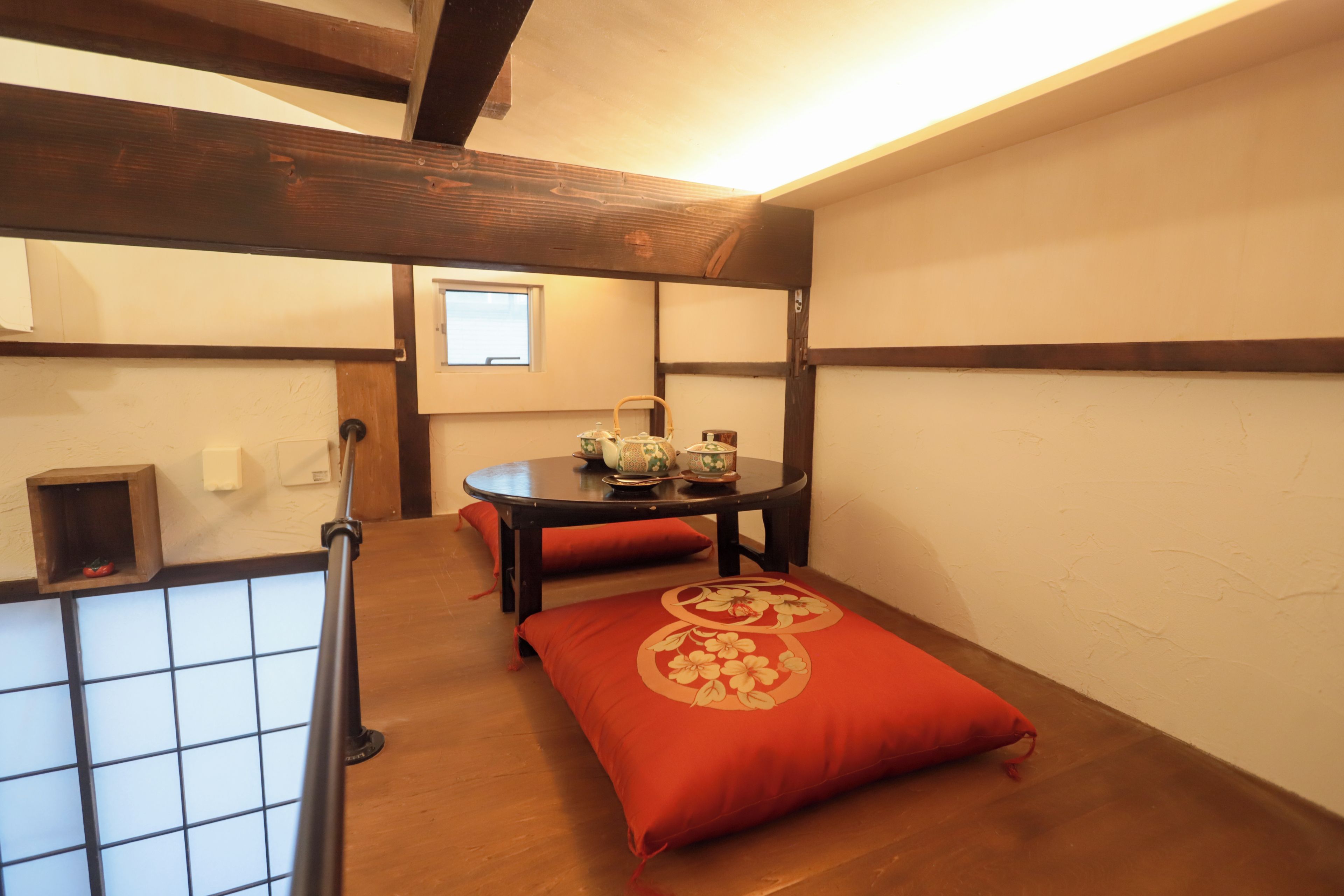 A room with a wooden floor, a table with a red cushion, a window, and a small shelf with a red object.