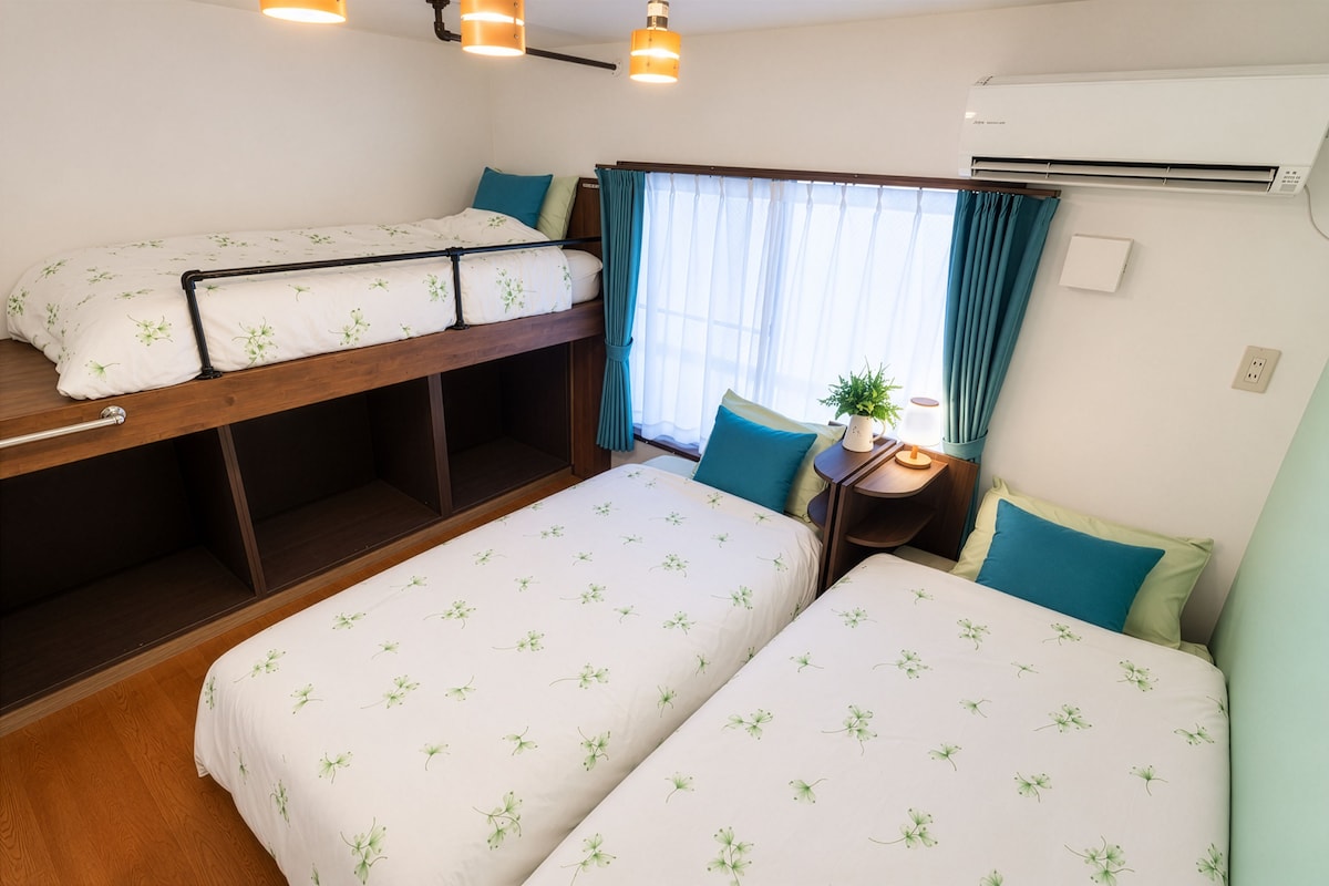Habitación de hotel con una cama litera, dos camas individuales, una mesita de noche con lámpara, una planta en maceta y un equipo de aire acondicionado.