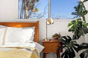 Quarto com cabeceira de madeira, cama com roupa de cama branca, mesa de cabeceira com abajur, uma pequena planta e uma grande planta Monstera.