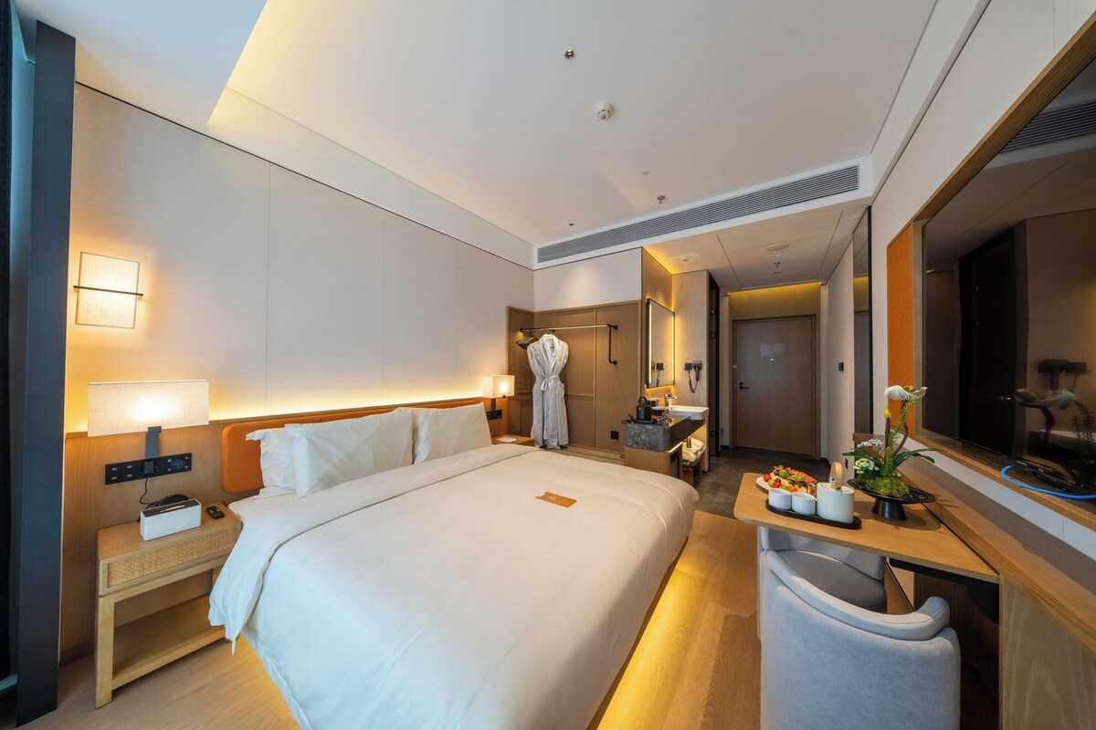 A hotel room with a large bed, bedside tables, a desk with a chair, a mirror, and a bathroom visible through an open door.