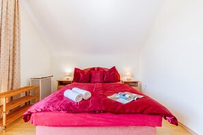 A single-bed room with a red bedspread, two pillows, a rolled towel, and a magazine on the bed. There is a wooden bench to the left and a small table with a lamp to the right.