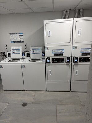 Eine Wäschemöglichkeit mit mehreren Waschmaschinen und Trocknern, einem Schwarzesbrett mit Informationen rund um die Wäsche und einem Lüftungssystem an der Decke.