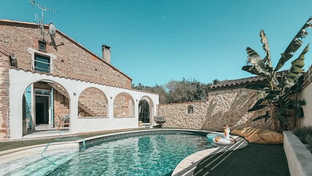 Une piscine à l’eau claire, un bâtiment en pierre doté d’arcades et un bananier.