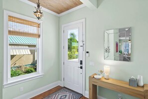 Ein Zimmer mit Tür, einem Fenster mit Jalousien, einem Spiegel, einer Deckenleuchte und einem Konsolentisch mit Lampe und Dekoration.