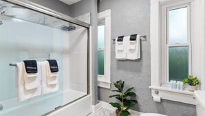 Een moderne badkamer met een douchecabine van glas, witte tegels, een potplant en een raam met handdoeken aan de wand.