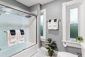 Ein modernes Badezimmer mit einer Duschkabine aus Glas, weißen Fliesen, einer Topfpflanze und einem Fenster, an dem Handtücher hängen.