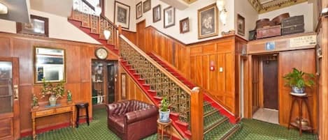 A hotel lobby with a staircase, a leather sofa, a small table with a plant, and a sign indicating the location of the toilets.
