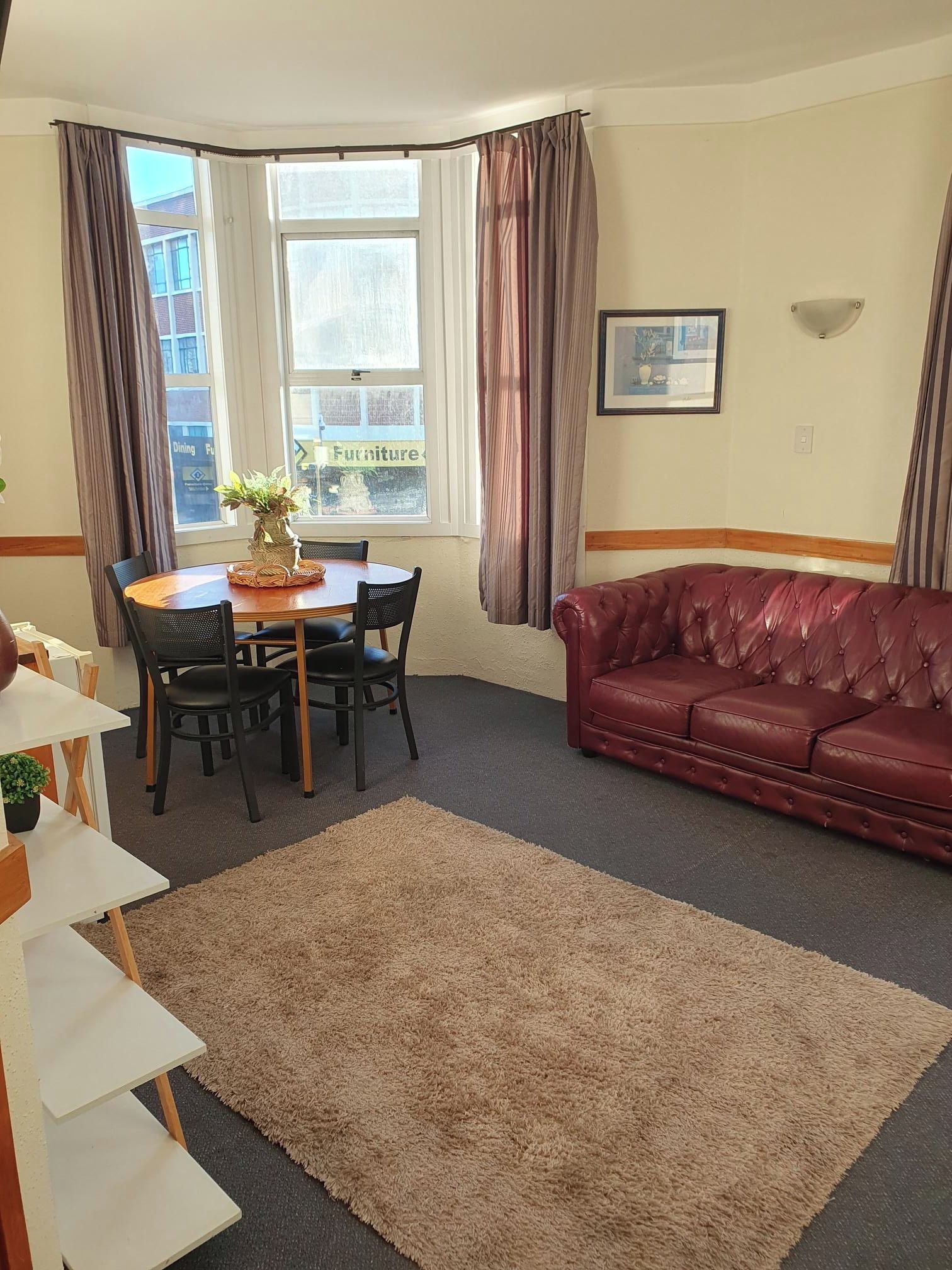 Ein möbliertes Wohnzimmer mit Esstisch, Stühlen, einem roten Ledersofa, einem großen Fenster mit Vorhängen und einem gerahmten Bild an der Wand.