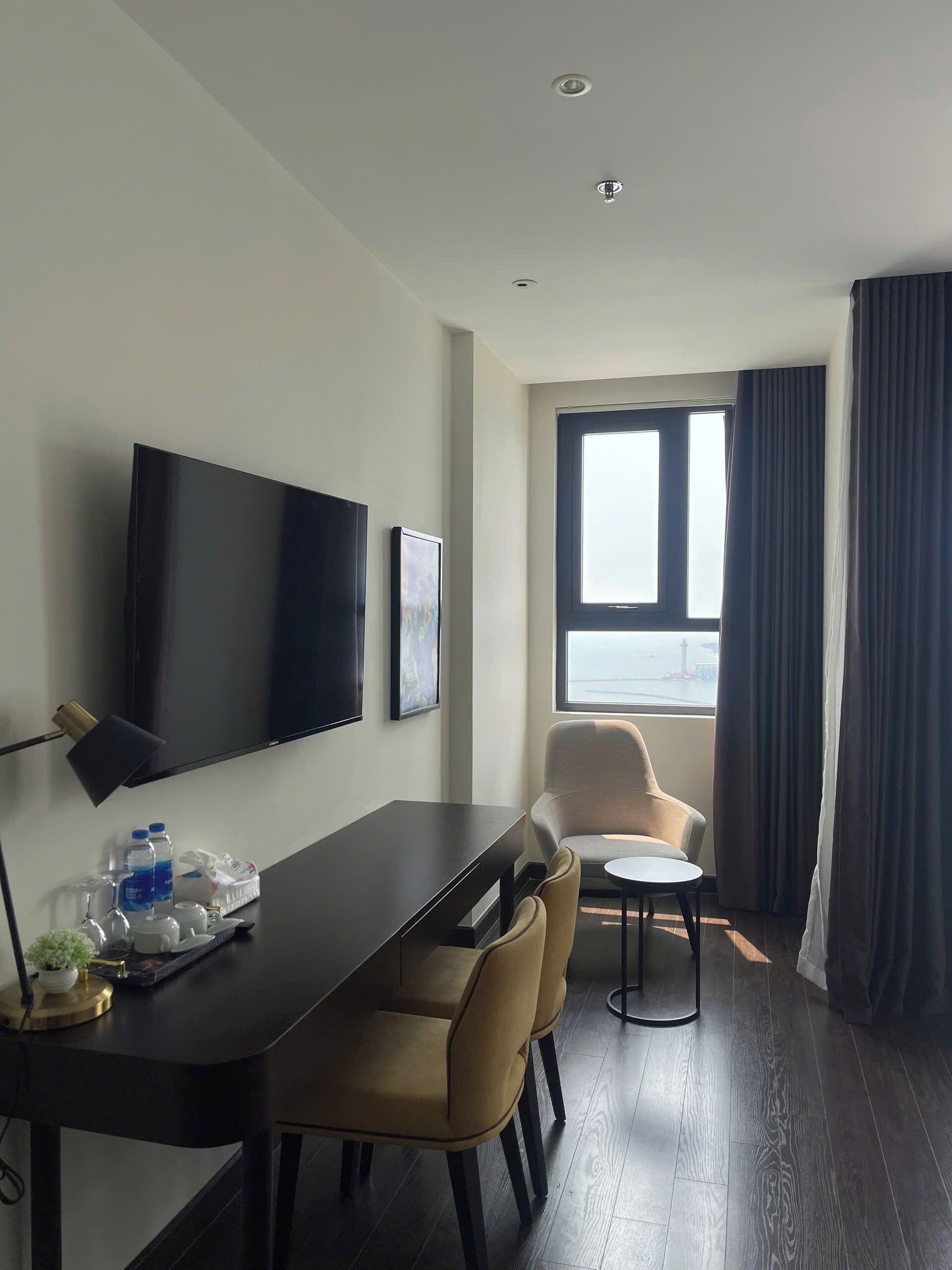 A hotel room with a flat-screen TV, a dining table with chairs, a small round table, a window with curtains, and a view of the cityscape.