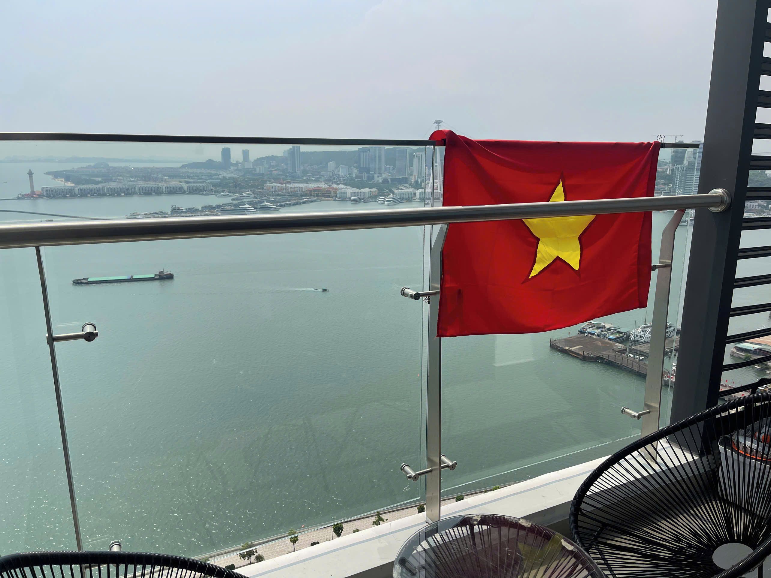 Un balcón con una bandera, vistas de un horizonte urbano y una masa de agua.