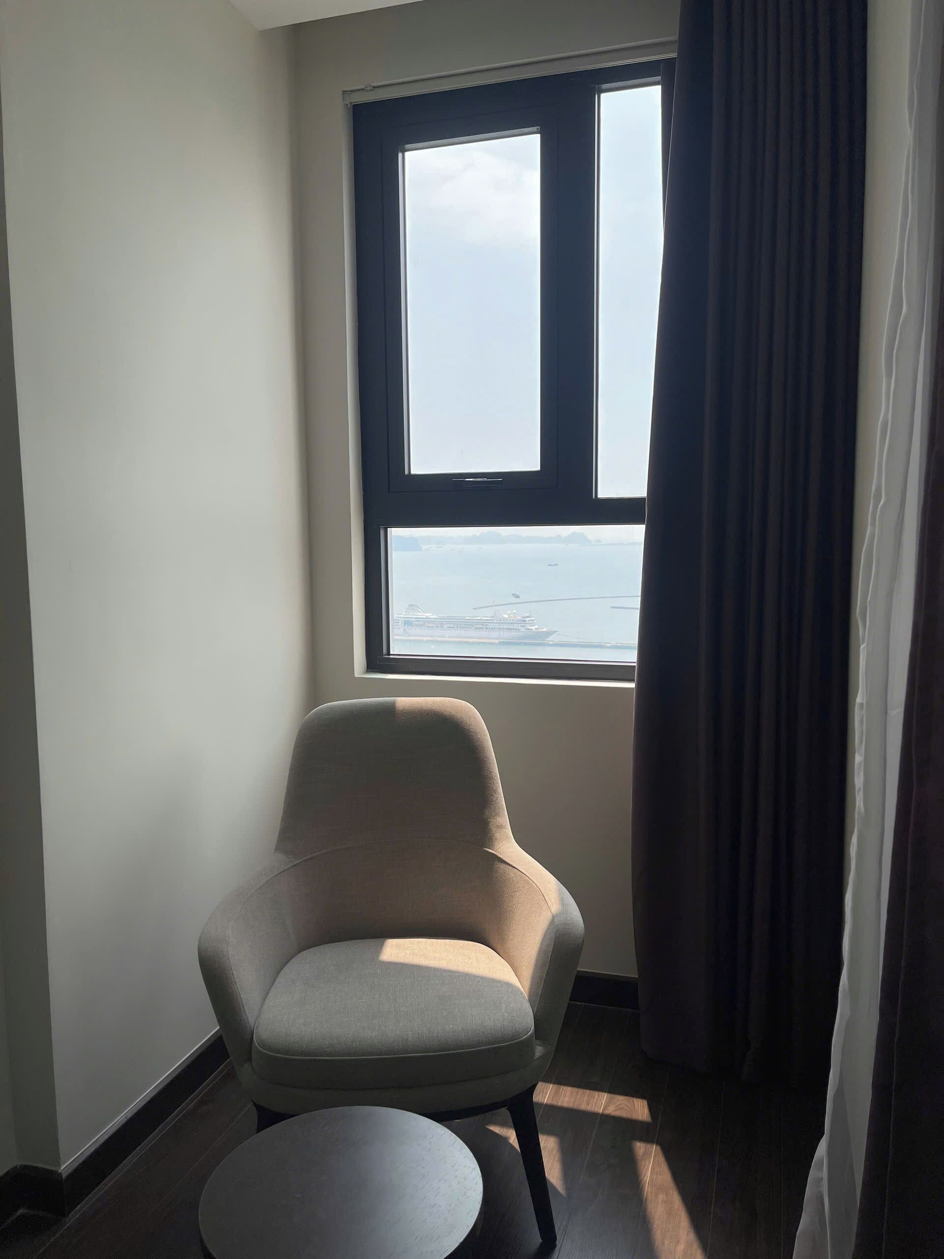 A room with a single chair and a table, a window with a view of the sea, and a curtain partially drawn.