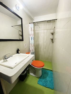 Un bany amb un lavabo blanc, un vàter amb el seient taronja, una cortina de dutxa i un tapet de bany blau.