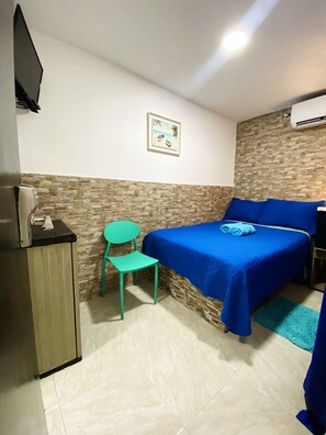 A room with a bed, a chair, a small cabinet, a picture on the wall, and a tiled floor.