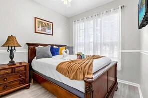 Un dormitorio con cama de madera, ropa de cama blanca, una lámpara de noche, un aparador y una ventana con persianas.