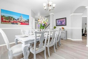 A dining area with a white table set for six, white chairs, a chandelier, and wall art depicting a castle. - 7BR Windsor Hills Heated Pool Game Room NearDisney (Kissimmee)