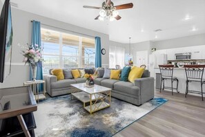 A modern living room with a grey sofa, a coffee table, and a dining area with a kitchen in the background. - 7BR Windsor Hills Heated Pool Game Room NearDisney (Kissimmee)
