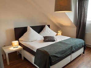 A neatly made bed with white linens and a dark green throw, a bedside lamp, and a small table with a lamp. - Hotel Zum Dorfbrunnen (Schleusingen)