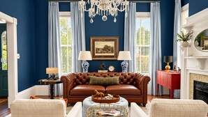 Une pièce comprenant un canapé en cuir marron, deux fauteuils de couleur crème, une table basse ronde, un lustre, une cheminée et un tableau encadré accroché au mur.