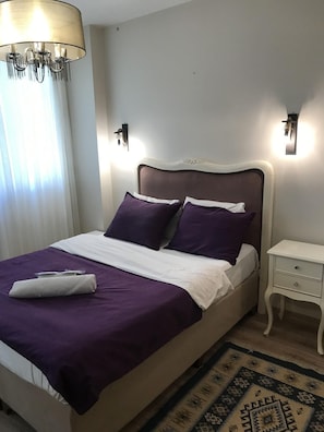 Siististi pedattu sänky, jossa on valkoinen ja violetti päiväpeite, valkoinen tyyny ja samanväriset peitot. Sängyn vieressä on valkoinen yöpöytä, jossa on yksi laatikko. Lattialla on kuvioitu matto ja seinälle on asennettu valaisin.