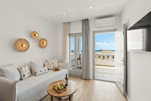 A modern living room with a sofa, a round wooden table with fruit and a bottle, and a balcony with a view of the sea. - Nikos & Maria Studios (Naxos)