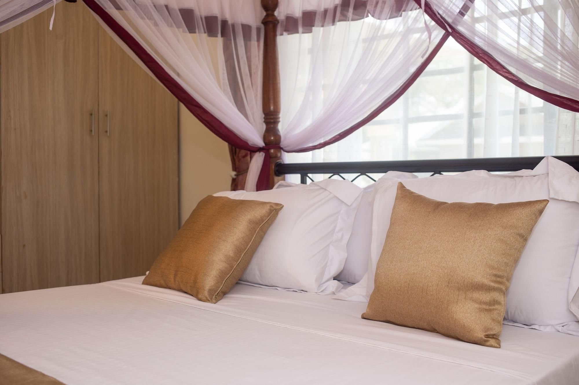A bed with white bedding and two gold pillows, a wooden headboard, and a window with sheer curtains.