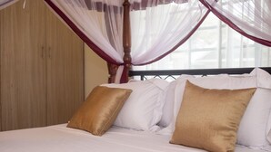 Ein Bett mit weißer Bettwäsche, zwei goldenen Kissen, einem Holz-Kopfteil und einem Fenster mit transparenten Vorhängen.