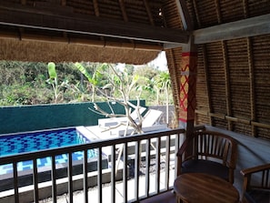 Área de piscina com deck de madeira, uma cadeira e estrutura com telhado de palha.