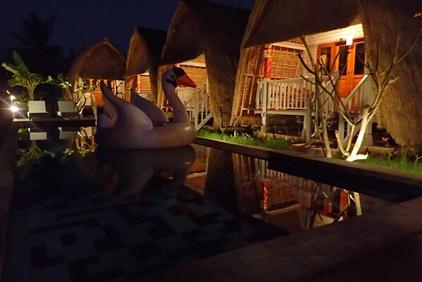 Vista noturna de um resort com bangalôs sobre a água, uma piscina e o reflexo dos bangalôs na água.