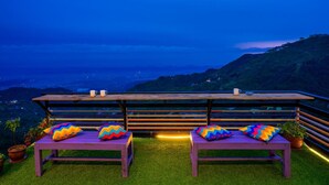 Terraço com duas bancos de madeira e almofadas coloridas, com vista para uma paisagem montanhosa ao entardecer.
