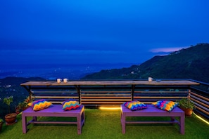 A terrace with two wooden benches and colorful cushions, overlooking a mountainous landscape at dusk. - Gostops Mussoorie (Dehradun)