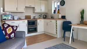 A compact kitchen with white cabinets, a microwave, a toaster, and a coffee maker. There is a dining area with a blue chair and a small table. - Waterside (Rye)