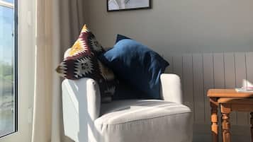 A corner room with a white armchair, a blue pillow, a patterned rug, a wooden table, and two framed botanical prints on the wall.