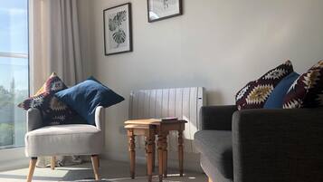 A living room with a grey sofa, a white armchair, a small wooden table, and a patterned rug. There are two framed pictures on the wall.