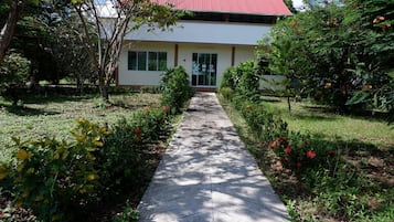 A pathway leading to a building with a red roof, surrounded by greenery and flowering plants.