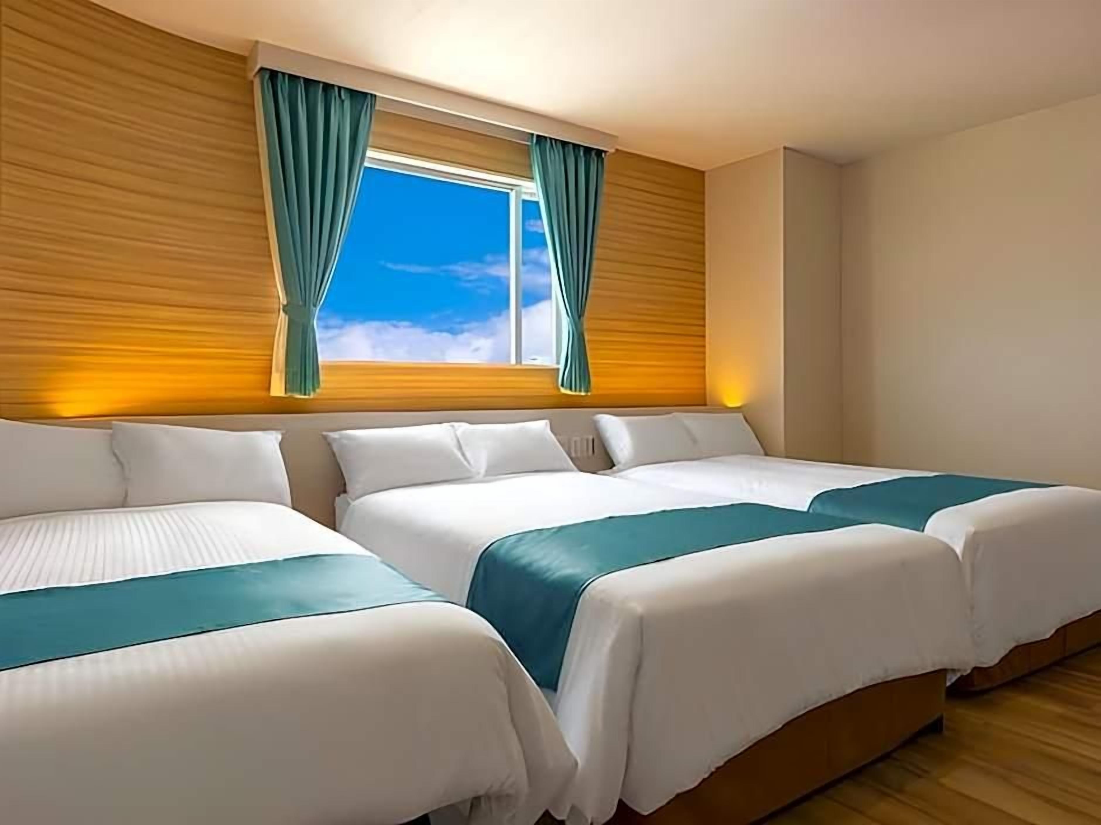 A hotel room with two beds, wooden paneling on the walls, a window with curtains, and a view of the sky.