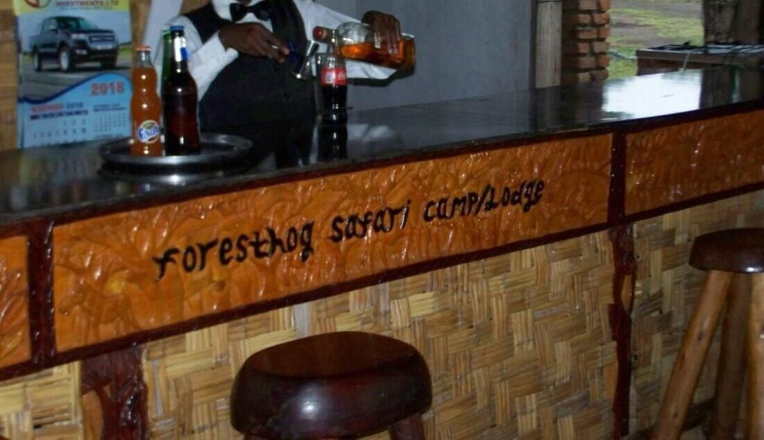 A bar area with a wooden counter and stools. The counter has a sign that reads 'forest safari camp lodge.' There are bottles and a glass on the counter.