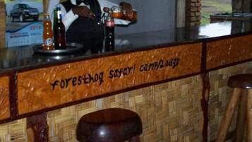 A bar area with a wooden counter and stools. The counter has a sign that reads 'forest safari camp lodge.' There are bottles and a glass on the counter.