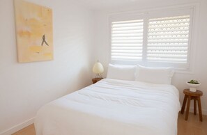 Ett sovrum med en säng, ett sängbord med en lampa, ett fönster med persienner och en tavla som hänger på väggen.