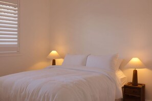 Ein Einzelbett mit weißer Bettwäsche, zwei Nachttische mit Lampen und ein Fenster mit Jalousien.