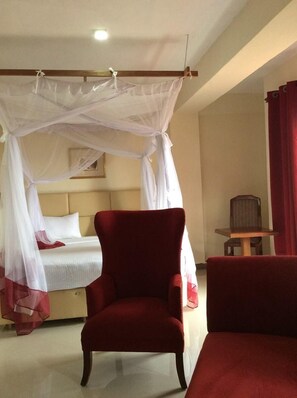 A hotel room with a bed, a red armchair, a wooden nightstand, and a red curtain. - Treat House Resort (Kisii)