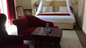 Ein Hotelzimmer mit einem Bett, einem Nachttisch mit einer Flasche, einem Sessel und einem Standventilator.