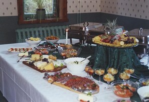 Una mesa de buffet con diferentes alimentos y bebidas, una planta en maceta y una ventana al fondo.