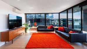 A modern living room with a large flat-screen TV, a sofa, a coffee table, and a vibrant orange rug. - The Yarra Terrace | 3BR with Pool & Parking (Abbotsford)