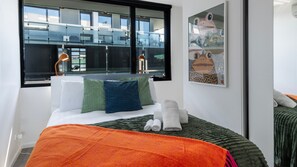 Een hotelkamer met een bed, een raam met uitzicht op gebouwen, een kunstwerk in een lijst aan de muur en een nachtkastje met lampen.
